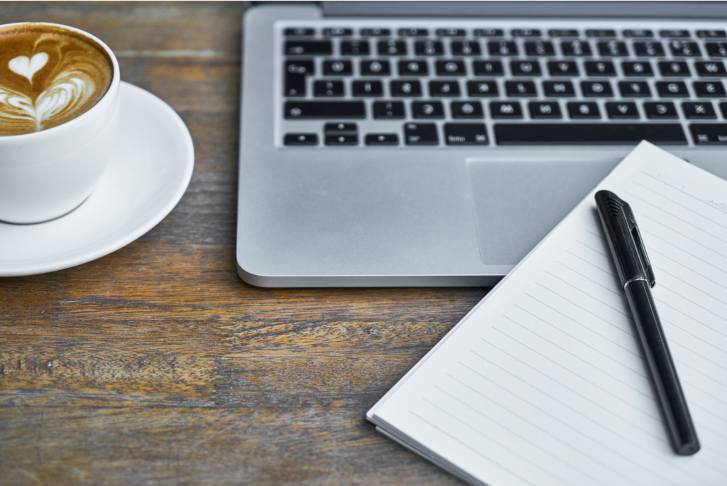 ordinateur portable ouvert sur une table en bois accompagné d'un cappuccino et d'un carnet de note ouvert avec un stylo