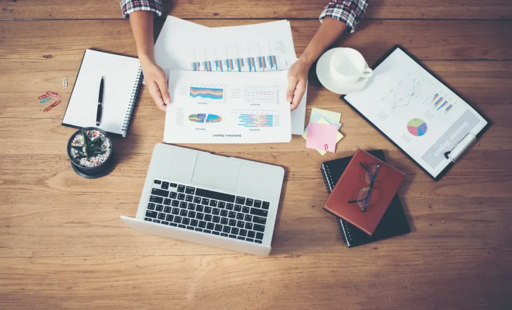 Les mains d'une femme tenant un document avec des graphiques et autour sur la table en bois un ordinateur, une tasse, un carnet avec stylo, et d'autres feuilles